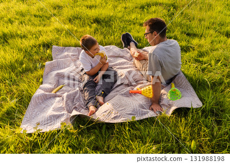 A father and his young son relax on a blanket in a lush green park. They share snacks and play with colorful toys, enjoying a sunny afternoon. Laughter fills the air as they bond A father and his young son relax on a blanket in a lush green park. They share snacks and play with colorful toys, enjoying a sunny afternoon. Laughter fills the air as they bond 131988198