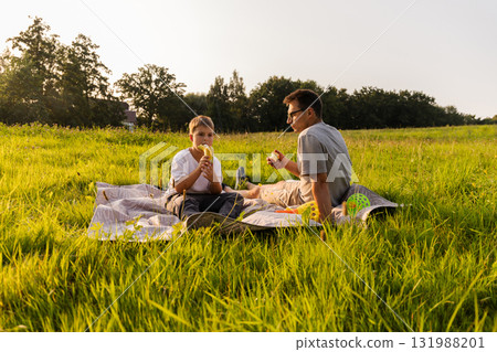 A father and son share a joyful afternoon together on a picnic blanket in a sunny field. They are eating apples, surrounded by green grass and trees, creating a warm family moment A father and son share a joyful afternoon together on a picnic blanket in a sunny field. They are eating apples, surrounded by green grass and trees, creating a warm family moment 131988201