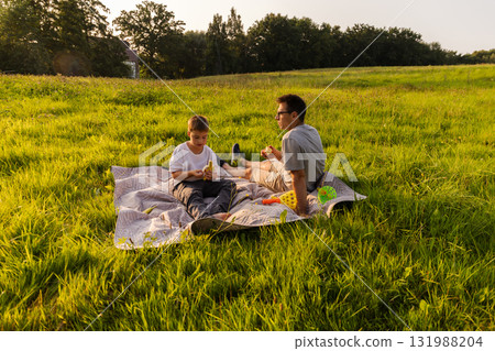 A father and his son sit on a blanket in a vibrant green field, sharing snacks and enjoying each other's company on a sunny afternoon 131988204