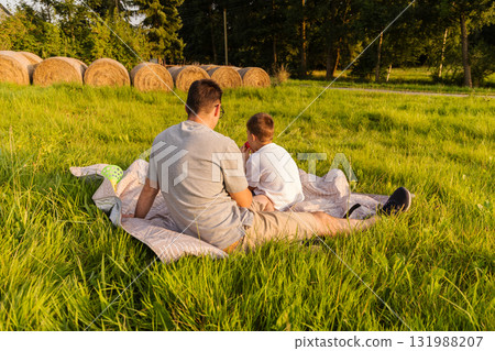 A father and son sit together on a blanket in a grassy field, enjoying quality time in nature. They appear relaxed as they share stories and laughter under the warm sun A father and son sit together on a blanket in a grassy field, enjoying quality time in nature. They appear relaxed as they share stories and laughter under the warm sun 131988207