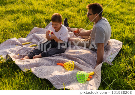 Spending quality time together, a father and son sit on a soft blanket in a vibrant green park. They snack and share laughter while enjoying nature's beauty on a sunny day 131988208