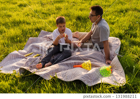 A father and son relax on a blanket in a lush green field, enjoying snacks together. The sun shines brightly as they bond over playful conversation and laughter 131988211