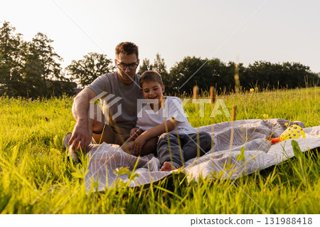 A father and his young son relax on a blanket in a vibrant green field. They are engaged in a fun activity while enjoying the warmth of the sun on a beautiful day 131988418