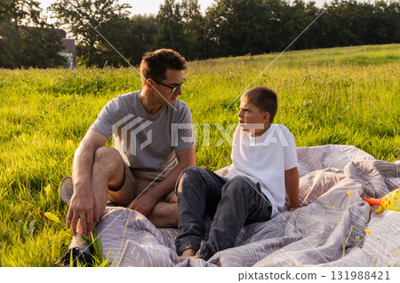 A father sits on a blanket in a grassy field, facing his son, who looks angry. They are spending time together in nature during late afternoon, surrounded by trees and open space A father sits on a blanket in a grassy field, facing his son, who looks angry. They are spending time together in nature during late afternoon, surrounded by trees and open space 131988421