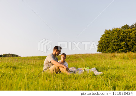 A father and son sit together on a blanket in a grassy field. They smile and talk while enjoying the warm sun and nature around them, creating happy memories A father and son sit together on a blanket in a grassy field. They smile and talk while enjoying the warm sun and nature around them, creating happy memories 131988424