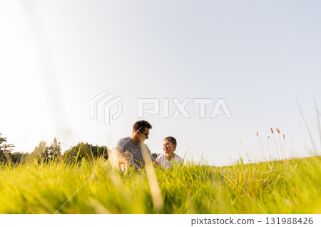 A father and his son sit in a lush green field, smiling and laughing together. They are enjoying a beautiful sunny day, embracing nature and each other's company 131988426