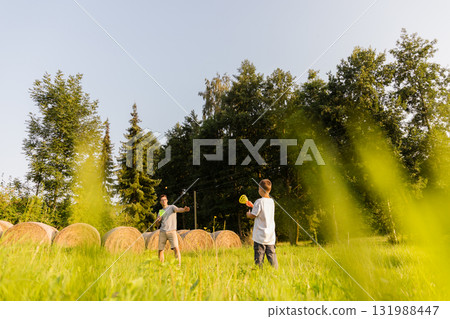 A father and son are joyfully playing catch in a lush green field under a clear sky. Hay bales line the background, creating a peaceful rural setting A father and son are joyfully playing catch in a lush green field under a clear sky. Hay bales line the background, creating a peaceful rural setting 131988447