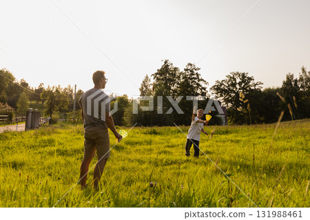 Under a clear sky, a father and son share a joyful moment playing catch in a green field. The sun shines brightly as they bond and have fun together in nature 131988461