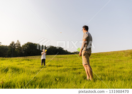 A father and son spend quality time together in a vibrant meadow. They are playing catch with a ball, surrounded by trees and grass, during a sunny afternoon 131988470