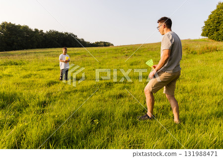 A father and son enjoy an afternoon outdoors, playing catch in a lush green field. The sun shines brightly as they engage in this joyful activity, surrounded by peaceful nature scenery 131988471