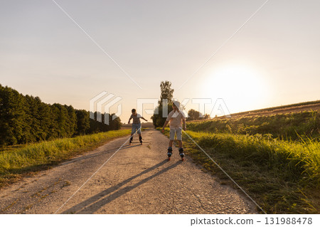 Two friends are having fun inline skating on a quiet path surrounded by greenery. The sun sets, creating a warm and bright atmosphere for outdoor activity 131988478