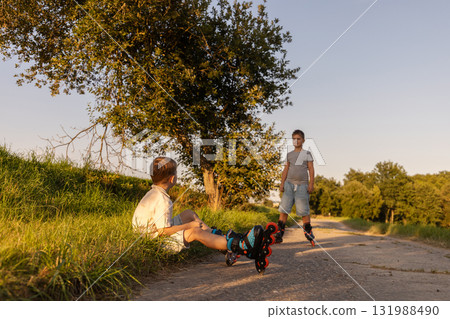Two friends are having fun inline skating on a path surrounded by greenery. One child sits on the ground while the other stands nearby, both enjoying their time outdoors 131988490
