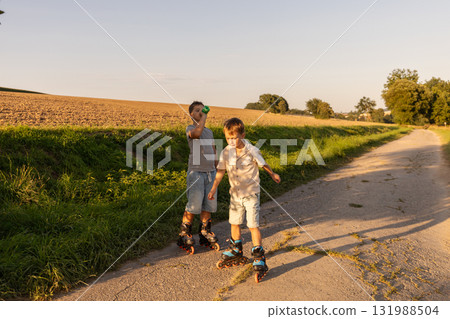 Two children skate on smooth pavement in a natural setting during a warm afternoon. They share laughter and fun as they glide alongside each other 131988504