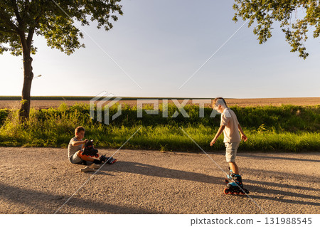Two friends are spending their free time inline skating on a quiet road surrounded by green grass and trees. It's a sunny afternoon with clear skies 131988545