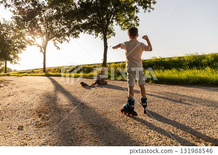 Two friends are outside roller skating along a sunny path in the park. One child is gliding forward while the other sits nearby, both enjoying their day in nature Two friends are outside roller skating along a sunny path in the park. One child is gliding forward while the other sits nearby, both enjoying their day in nature 131988546