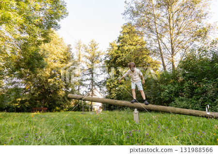 Two children are playing outdoors in a park, balancing on a wooden log. Surrounded by tall trees and lush grass, they are enjoying a sunny day filled with laughter and activity Two children are playing outdoors in a park, balancing on a wooden log. Surrounded by tall trees and lush grass, they are enjoying a sunny day filled with laughter and activity 131988566