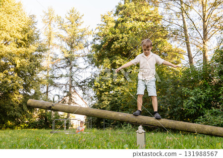Two children are playing outside in a lush park. One child balances on a log while the other plays nearby, enjoying the sunny weather and the beauty of nature Two children are playing outside in a lush park. One child balances on a log while the other plays nearby, enjoying the sunny weather and the beauty of nature 131988567