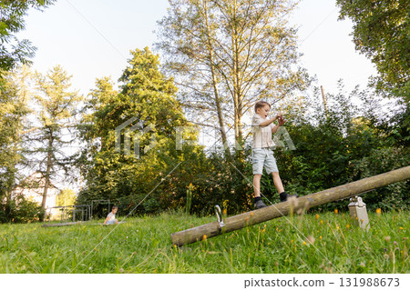Two children engage in playful activities in a lush green park during daylight. One child balances on a log, while the other enjoys the surrounding nature 131988673