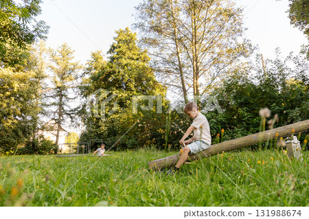 Two children are playing outdoors in a lush green park filled with trees. One child balances on a log while the other enjoys the spacious area, making the most of their playful afternoon Two children are playing outdoors in a lush green park filled with trees. One child balances on a log while the other enjoys the spacious area, making the most of their playful afternoon 131988674