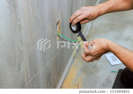 Hands working on wiring inside wall, carefully using tape to secure connections in renovated room setting. Hands working on wiring inside wall, carefully using tape to secure connections in renovated room setting. 131989801