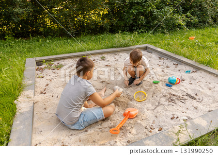 Two kids are playing in a sand pit surrounded by greenery. They are using colorful toys to build structures and enjoy each other's company on a sunny day Two kids are playing in a sand pit surrounded by greenery. They are using colorful toys to build structures and enjoy each other's company on a sunny day 131990250