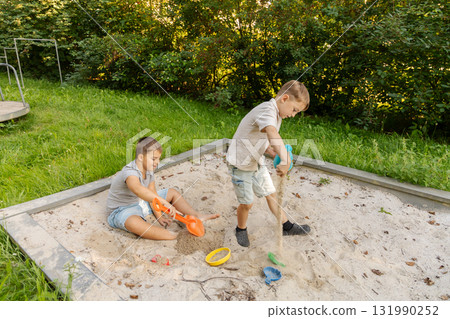 Two children are happily playing in a sandbox, digging with colorful shovels. Lush green grass and trees create a natural backdrop in a warm afternoon setting, enhancing their fun Two children are happily playing in a sandbox, digging with colorful shovels. Lush green grass and trees create a natural backdrop in a warm afternoon setting, enhancing their fun 131990252