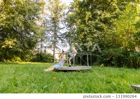 Two kids are having fun on a merry-go-round in a lush park. The sun shines brightly as they laugh and play, surrounded by tall trees and green grass 131990264
