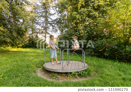 Two children are having fun on a merry-go-round in a lush park. They are smiling and enjoying nature while playing in the soft grass on a sunny day Two children are having fun on a merry-go-round in a lush park. They are smiling and enjoying nature while playing in the soft grass on a sunny day 131990270