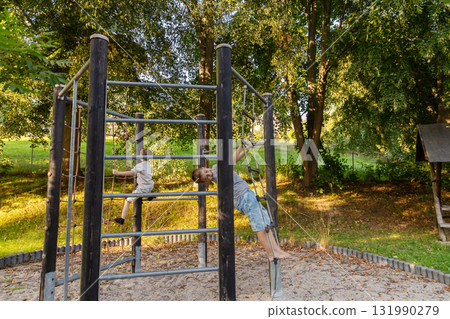 Two kids are having fun outdoors, climbing on a play structure in a park. They enjoy the fresh air and sunshine while exploring and playing together among the trees 131990279