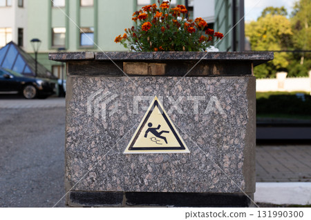 Cautionary slip hazard sign, affixed to a stone pillar with vibrant flowers on top. Reminder of safety in public spaces, promoting awareness of potential risks 131990300