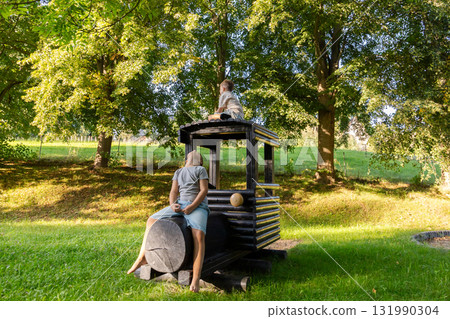 Two children are joyfully playing on a wooden train structure in a lush green park. They are immersed in their imaginative play surrounded by trees and sunlight Two children are joyfully playing on a wooden train structure in a lush green park. They are immersed in their imaginative play surrounded by trees and sunlight 131990304