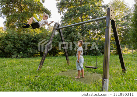 Two kids are playing joyfully in a green park. One child swings high while the other watches with excitement. Bright sunshine filters through the trees, creating a lively scene 131990311