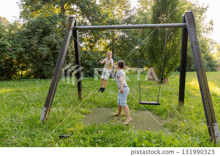 Two children are happily playing on swings in a vibrant park. The scene is filled with lush green grass and trees, radiating a joyful summer vibe 131990323