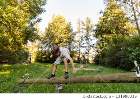 Two children are playing outside in a green park. One child balances on a seesaw while the other watches from a distance. The sun is shining brightly in the background 131990339