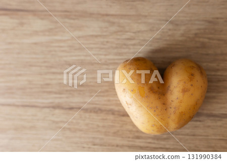 A unique heart-shaped potato rests on a smooth wooden table, showcasing its distinct shape and texture. The simple setting adds warmth, making it an intriguing sight for food lovers 131990384