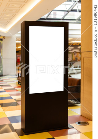 A tall digital display stand is placed indoors at a vibrant shopping mall. The screen is blank, presenting an opportunity for future advertising A tall digital display stand is placed indoors at a vibrant shopping mall. The screen is blank, presenting an opportunity for future advertising 131990442