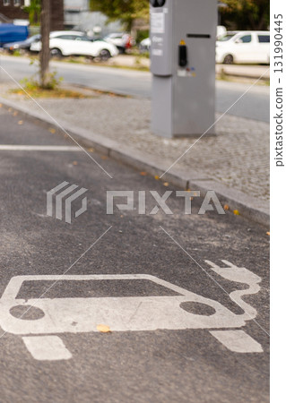 A clear marking for an electric vehicle charging station is visible on the pavement. The area is part of a city street, with parked cars and trees nearby, indicating a busy urban environment A clear marking for an electric vehicle charging station is visible on the pavement. The area is part of a city street, with parked cars and trees nearby, indicating a busy urban environment 131990445