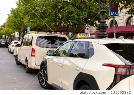 Taxis sit patiently in a line on a city street, waiting for passengers. Trees provide shade, and buildings line the avenue, creating a bustling urban atmosphere Taxis sit patiently in a line on a city street, waiting for passengers. Trees provide shade, and buildings line the avenue, creating a bustling urban atmosphere 131990588