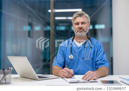 Male doctor in scrubs with stethoscope at a desk, writing on a clipboard while looking at the camera, conveying experience, focus and trustworthy patient care in a modern clinic 131990657
