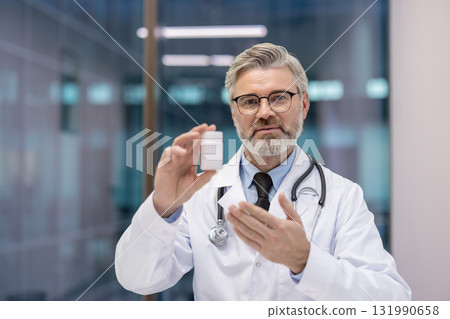 Senior male doctor wearing white lab coat and stethoscope, holding a blank white medicine bottle, and gesturing with his hand while explaining treatment options Senior male doctor wearing white lab coat and stethoscope, holding a blank white medicine bottle, and gesturing with his hand while explaining treatment options 131990658