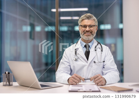Experienced male doctor in white coat with stethoscope sits at clinic desk with laptop and documents, smiling confidently at the camera conveying trustworthy medical care 131990679