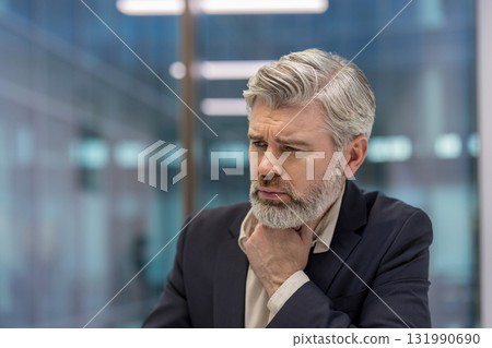 Senior businessman in suit touching his throat and wincing, showing sore throat and fatigue while working in a modern office, stressed and looking away, close-up headshot Senior businessman in suit touching his throat and wincing, showing sore throat and fatigue while working in a modern office, stressed and looking away, close-up headshot 131990690