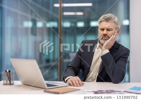 Mature experienced businessman sick at workplace, man suffering from toothache, holding hand to face Mature experienced businessman sick at workplace, man suffering from toothache, holding hand to face 131990702