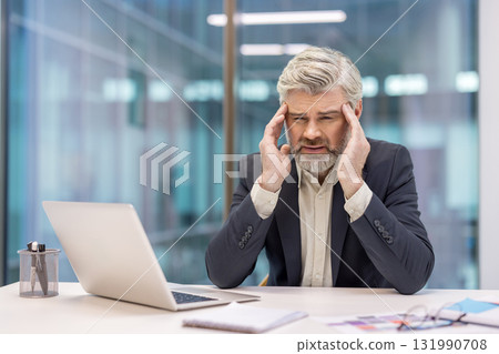 Senior businessman sitting at a desk in a modern office, grappling with a severe headache and expressing physical pain while working on a laptop, indicating burnout and job-related pressure Senior businessman sitting at a desk in a modern office, grappling with a severe headache and expressing physical pain while working on a laptop, indicating burnout and job-related pressure 131990708