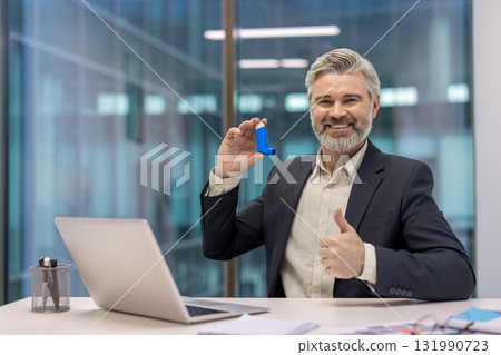 Smiling mature businessman sitting at his office desk, holding an asthma inhaler and giving a thumbs up, symbolizing health, medication, and managing chronic conditions in a professional environment Smiling mature businessman sitting at his office desk, holding an asthma inhaler and giving a thumbs up, symbolizing health, medication, and managing chronic conditions in a professional environment 131990723