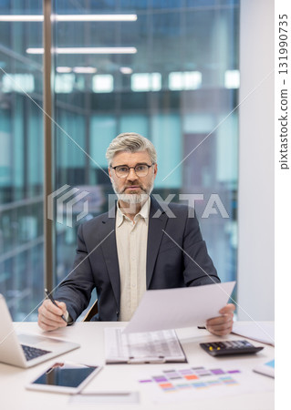 Senior businessman with gray beard and glasses reviews documents at a sleek corporate desk, pen in hand, laptop and calculator nearby, focused on strategy and financial planning Senior businessman with gray beard and glasses reviews documents at a sleek corporate desk, pen in hand, laptop and calculator nearby, focused on strategy and financial planning 131990735