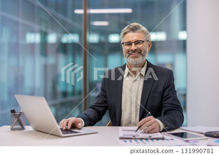Happy mature businessman working on laptop and reviewing documents at a desk in a contemporary office, representing success, professionalism, and experienced leadership in a corporate environment 131990781