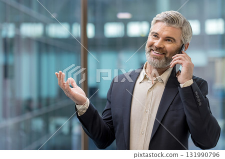 Mature businessman smiling and gesturing during a phone call, standing in an office with modern architecture, representing successful communication and corporate networking 131990796