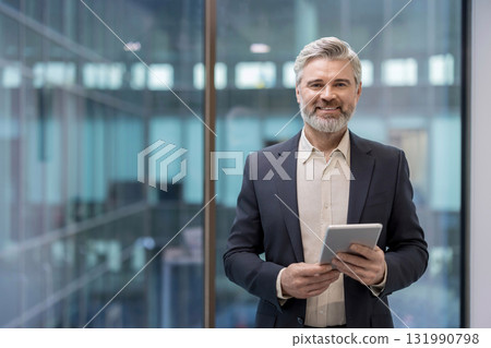 A smiling, distinguished businessman stands confidently holding a tablet in a modern office setting with large windows. A smiling, distinguished businessman stands confidently holding a tablet in a modern office setting with large windows. 131990798