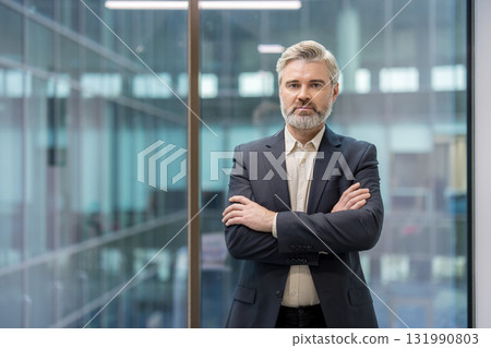 Mature businessman with gray hair and beard in a dark suit standing confidently with crossed arms. Looking at the camera against a modern office background. Symbolizing leadership and professionalism 131990803